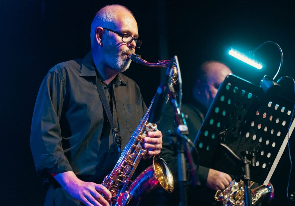 a lovely performance photograph showing a saxophonist in action during what appears to be a jazz performance. The musician is wearing glasses and a dark shirt, playing what looks like an alto or tenor saxophone. The atmospheric lighting creates a beautiful blue and purple ambiance typical of jazz club settings, with dramatic stage lighting highlighting the performer against the darker background.