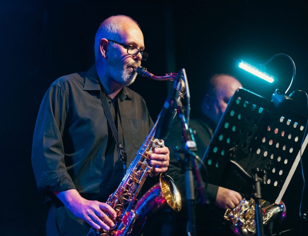 a lovely performance photograph showing a saxophonist in action during what appears to be a jazz performance. The musician is wearing glasses and a dark shirt, playing what looks like an alto or tenor saxophone. The atmospheric lighting creates a beautiful blue and purple ambiance typical of jazz club settings, with dramatic stage lighting highlighting the performer against the darker background.