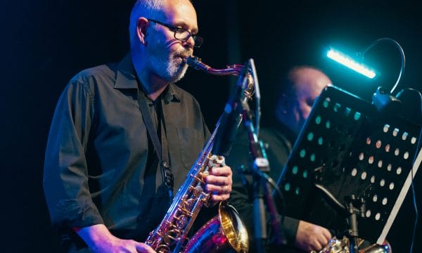 a lovely performance photograph showing a saxophonist in action during what appears to be a jazz performance. The musician is wearing glasses and a dark shirt, playing what looks like an alto or tenor saxophone. The atmospheric lighting creates a beautiful blue and purple ambiance typical of jazz club settings, with dramatic stage lighting highlighting the performer against the darker background.