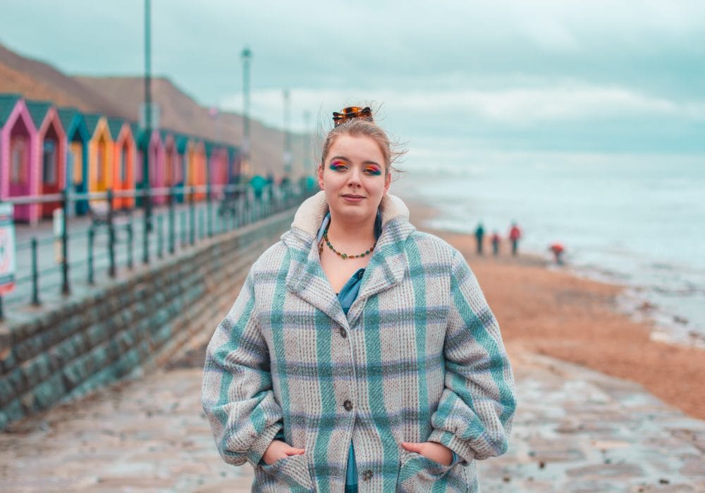 Lizzie Lovejoy stands on Saltburn beach (with the brightly coloured beach huts in the background). They are wearing a checked coat, bright eyeshadow, and their hair is tied up in a bun.