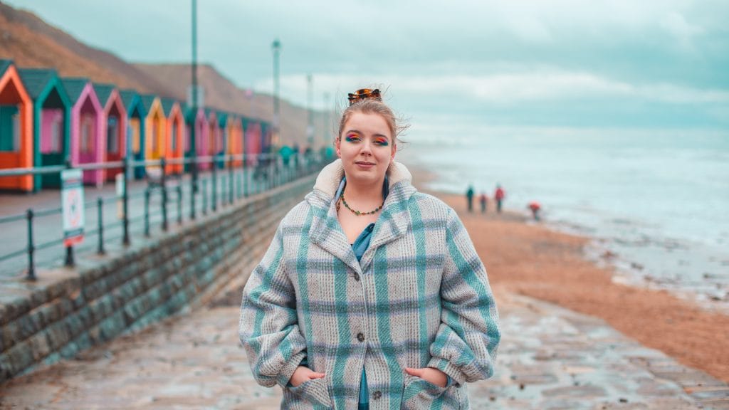 Lizzie Lovejoy stands on Saltburn beach (with the brightly coloured beach huts in the background). They are wearing a checked coat, bright eyeshadow, and their hair is tied up in a bun.