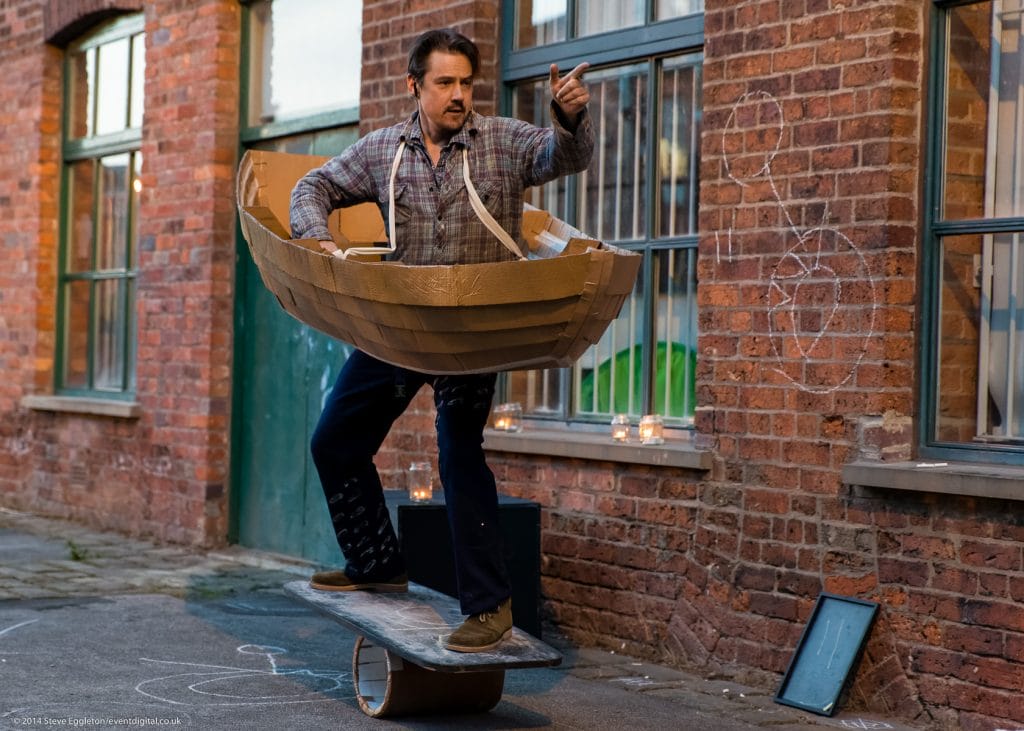 A man stands on a board balanced on a small barrel shaped object. He is wearing a cardboard boat hanging by straps over his shoulders.