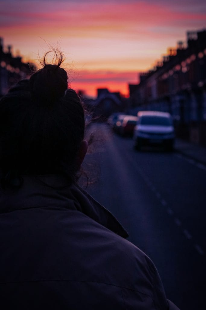 A figure in shadow in the foreground looks down a street lined with terraced houses and parked cars, towards a pink and orange sky.