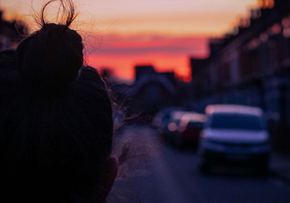 A figure in shadow in the foreground looks down a street lined with terraced houses and parked cars, towards a pink and orange sky.