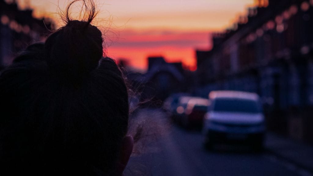 A figure in shadow in the foreground looks down a street lined with terraced houses and parked cars, towards a pink and orange sky.