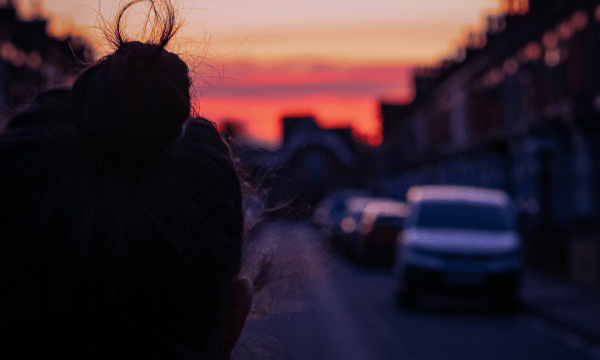 A figure in shadow in the foreground looks down a street lined with terraced houses and parked cars, towards a pink and orange sky.