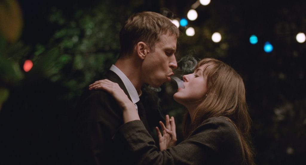 A man and woman stand very close facing each other, the man is blowing cigarette smoke into the woman's mouth