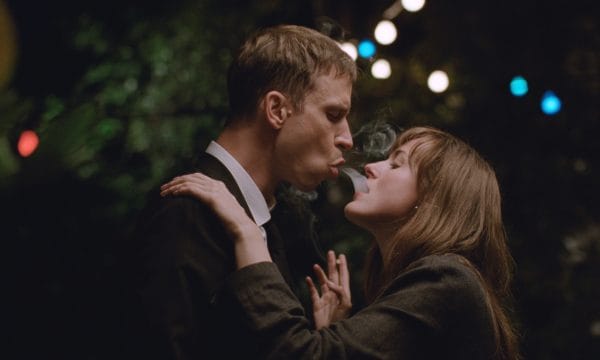 A man and woman stand very close facing each other, the man is blowing cigarette smoke into the woman's mouth