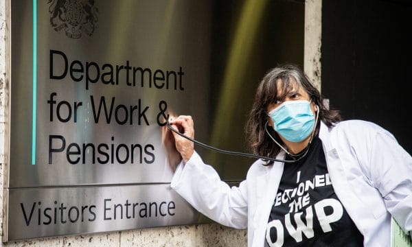 A person dressed in a doctor's coat and wearing a face mask holds a stethoscope to a metal sign on a wall reading 'Department for Work & Pensions / Visitors Entrance'