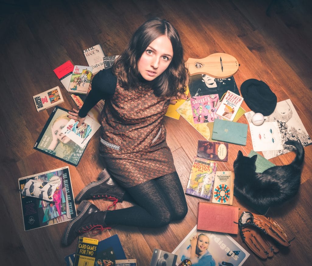 Singer Amelia Coburn sits on the floor looking up into the camera, she is surrounded by vinyl records.