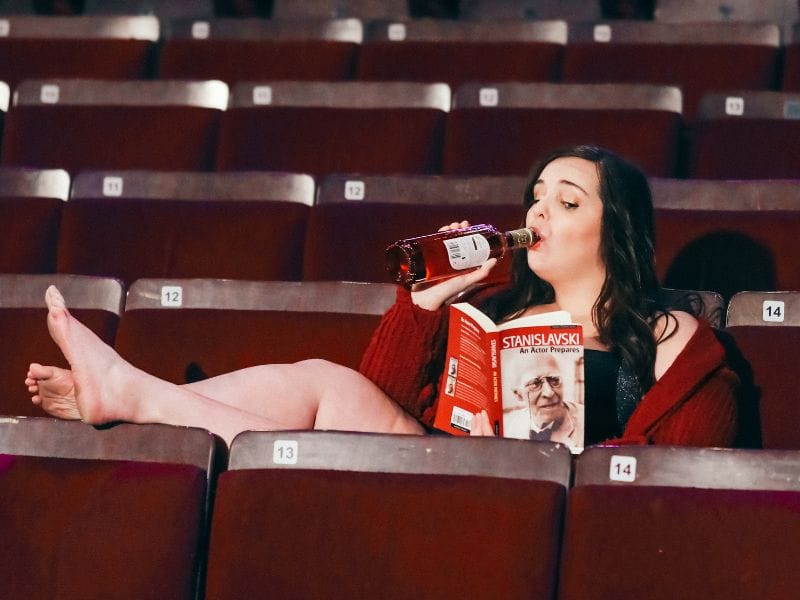 Rachel sits in an empty theatre, with her legs up on a seat in front of her, and a book in one hand and a bottle of wine in the other.