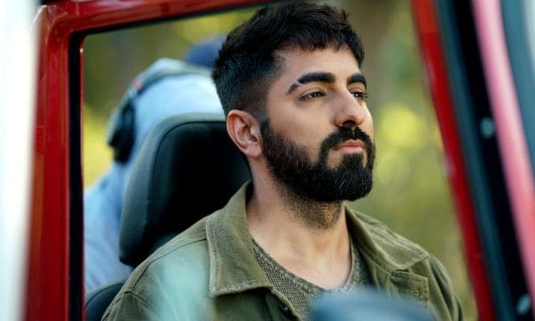 A south Asian man with dark hair and a beard sits in the front seat of a car, looking thoughtfully into the distance.