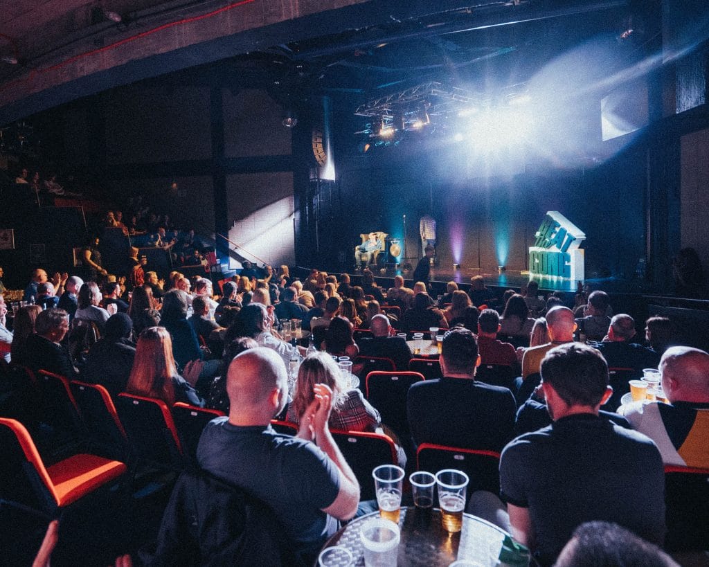 A crowded room lit by bright white stage lights, the audience are all laughing and clapping, on the stage is a 3d prop which reads Beat the Gong.