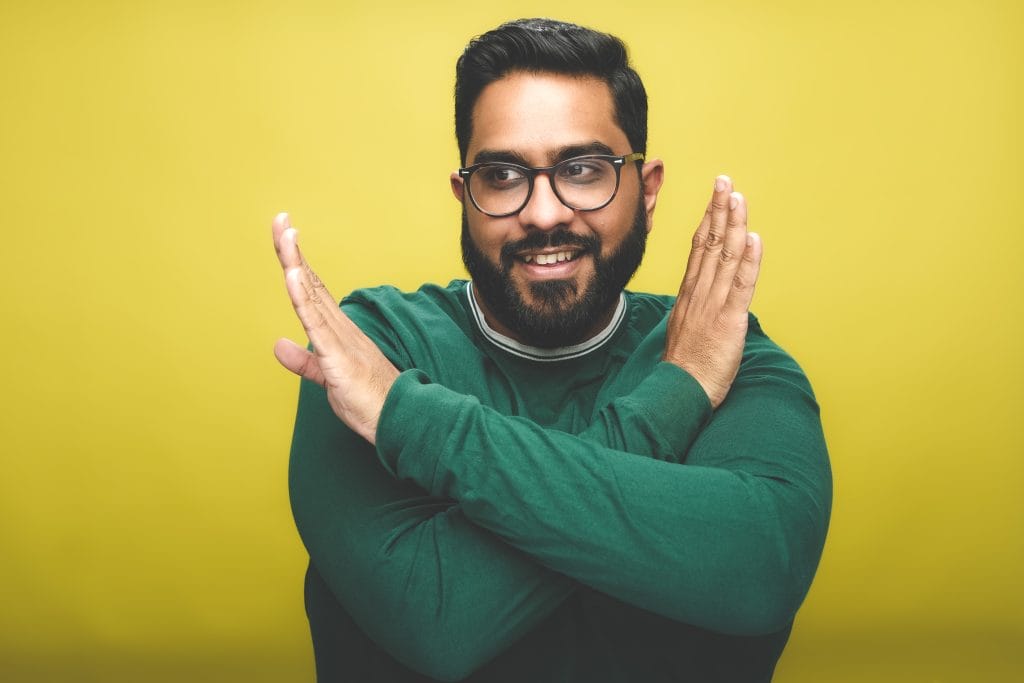 Comedian Eshaan Akbar stands in front of a yellow backdrop. He wears a green jumper, and has both of his arms across his body to make an X shape, he is looking off to the side of the camera and is smiling.