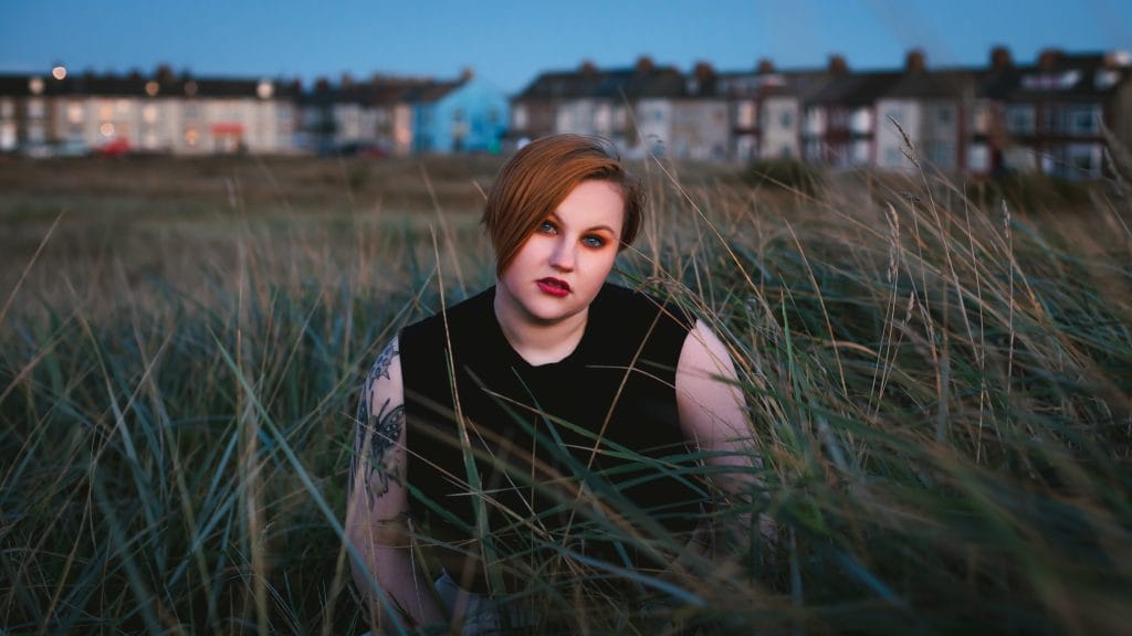 Artist Audrey Cook sits in a patch of long grass. There is a row of terraced houses against a dark blue sky in the distance behind them.