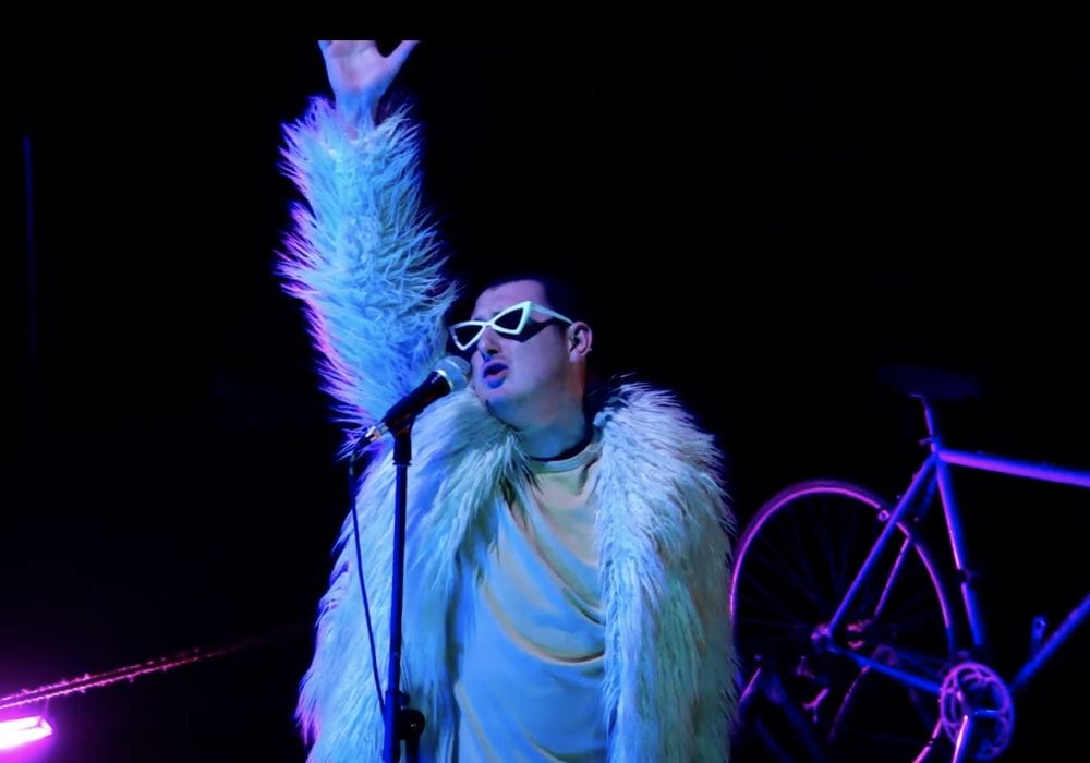 A person wearing sunglasses on a dark stage. A bicycle is behind them.