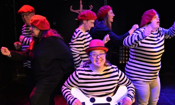 A female actor seated on a chair at the front smiling broadly holding a giant game controller. Five other actors in stripy white and black tops and red berets try to run off in all directions.