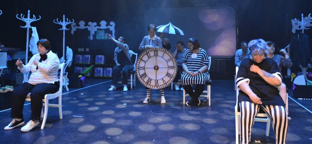 A scene from dress rehearsals. Most of the actors are seated and one is standing, holding a clock face with Roman numerals on it. All the actors are dressed in black and white, and there is a black and white striped umbrella at the back of the set, with white hat stands along each side.