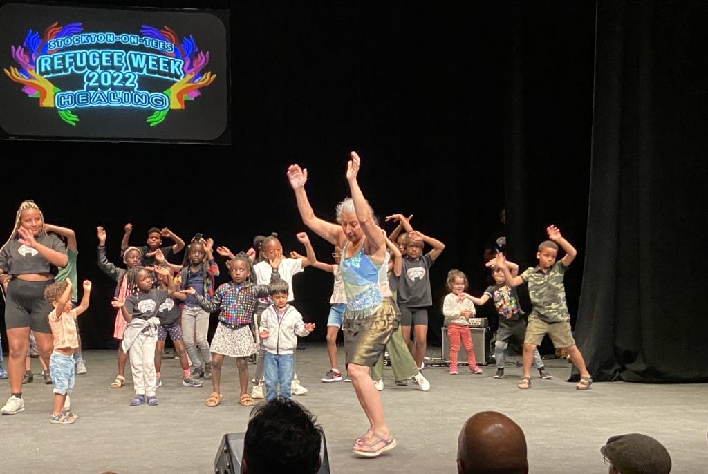 A group of people of all ages on a stage, they are all dancing to the left of the stage, they have their arms raised. On a screen behind them are the words Refugee Celebration 2022