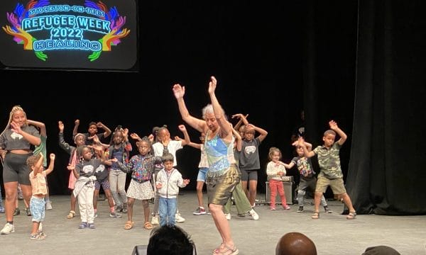 A group of people of all ages on a stage, they are all dancing to the left of the stage, they have their arms raised. On a screen behind them are the words Refugee Celebration 2022