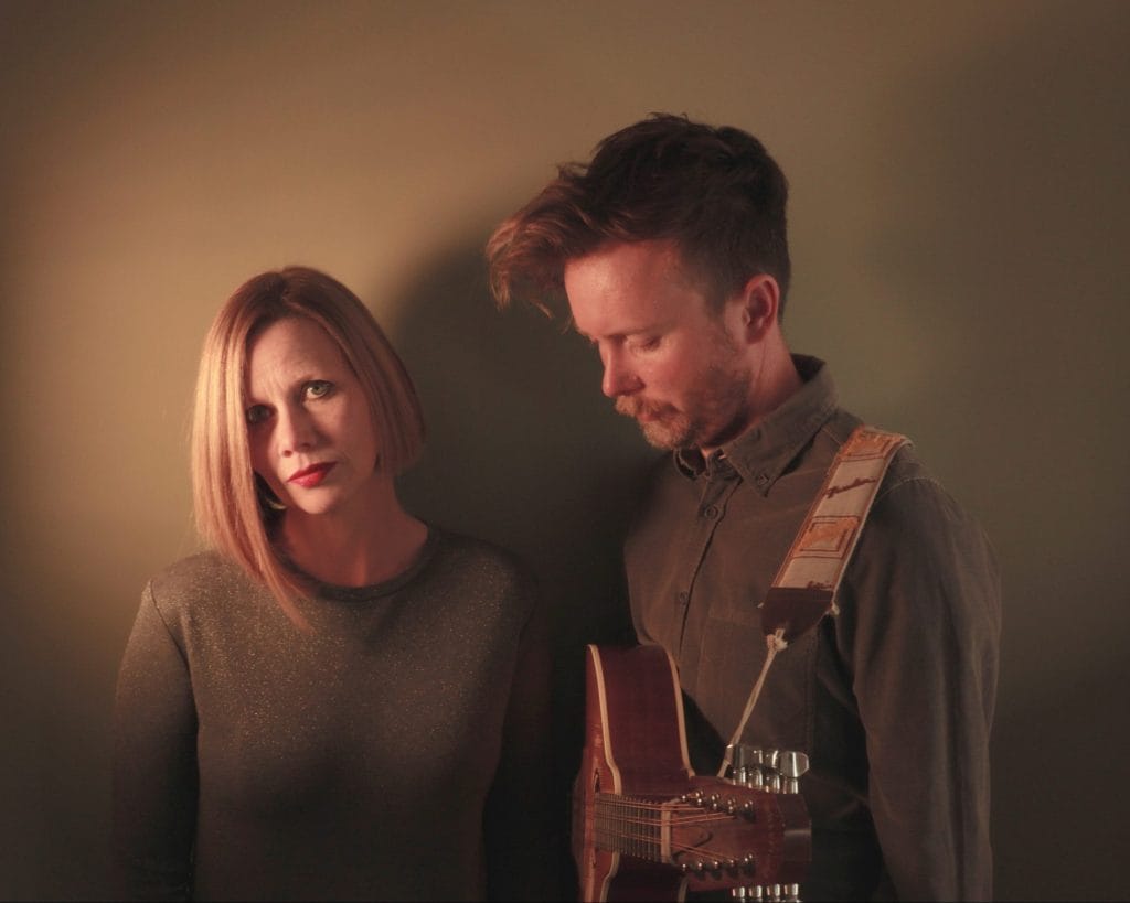 The band Megson stand in front of a dimly lit backdrop. On the left, a blonde female presenting person stares into the camera, and on the right a male presenting person holding a guitar looks down at the ground.