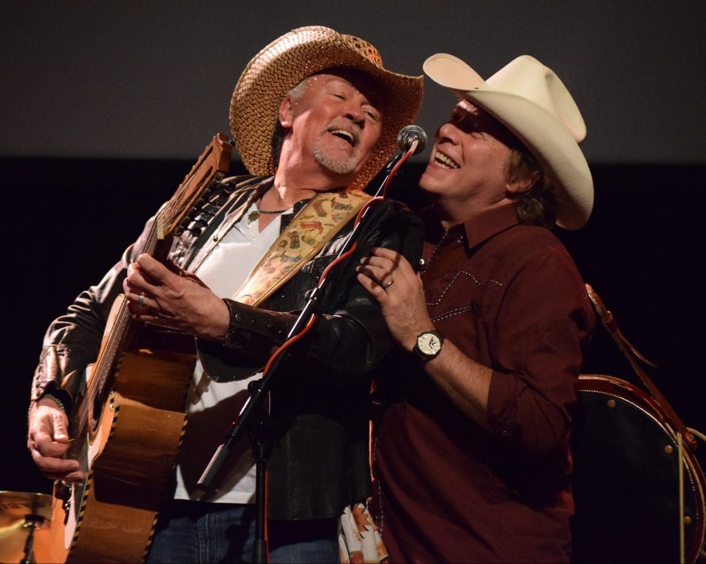 Two male musicians are both singing into a microphone, one man holds an acoustic guitar, both are wearing cowboy hats.