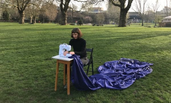 Natasha sits outside in an open, grassed area. She is sitting on a chair using a white sewing machine on a small table in front of her. A swathe of dark blue material fans out behind her from the sewing machine.