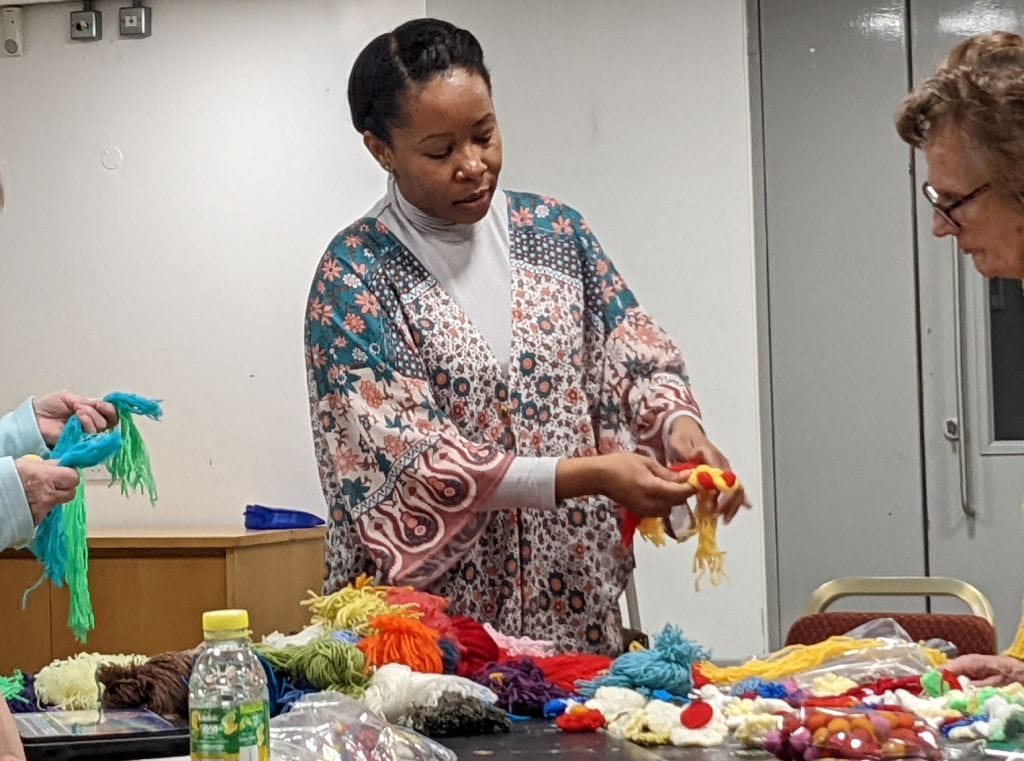 A photo of artist Jane, she is displaying some recycled wool jewelry to a participant. The table in front of her is covered in multicoloured balls of wool. Just out of shot, another participant is holding their own wool necklace made from blue and green wool.