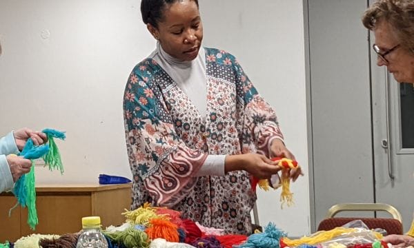 A photo of artist Jane, she is displaying some recycled wool jewelry to a class participant. The table in front of her is covered in multicoloured balls of wool. Just out of shot, another class participant is holding their own wool necklace made from blue and green wool.