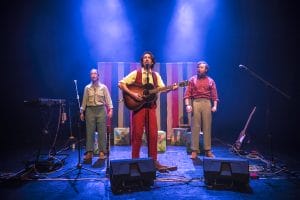 One performer behind a microphone with a guitar and two other performers behind him to either side