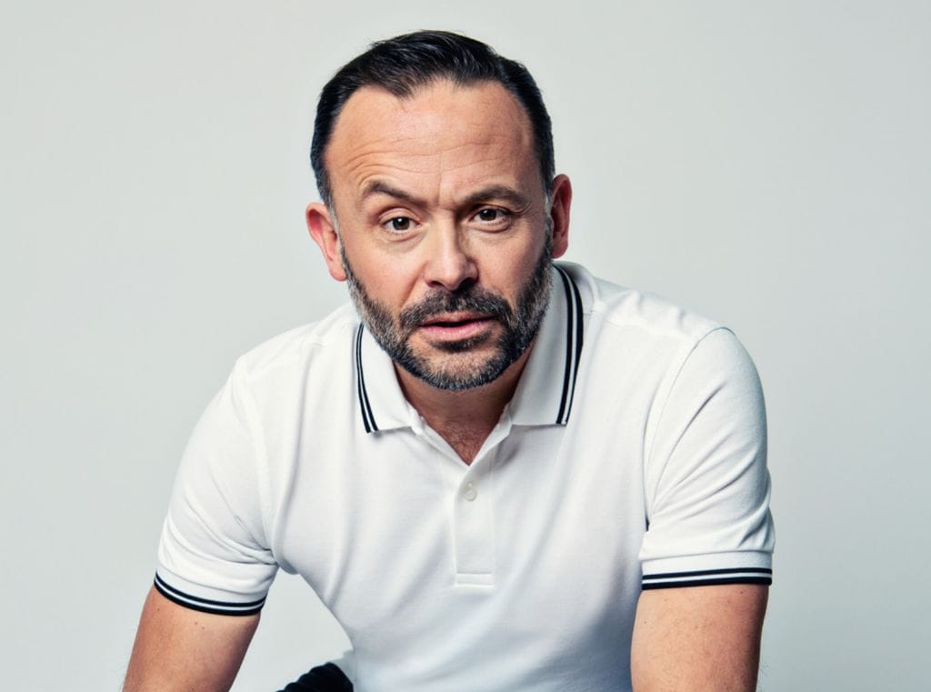 A headshot of comedian Geoff Norcott, he is wearing a white polo shirt and is looking intently into the camera with one eyebrow raised.