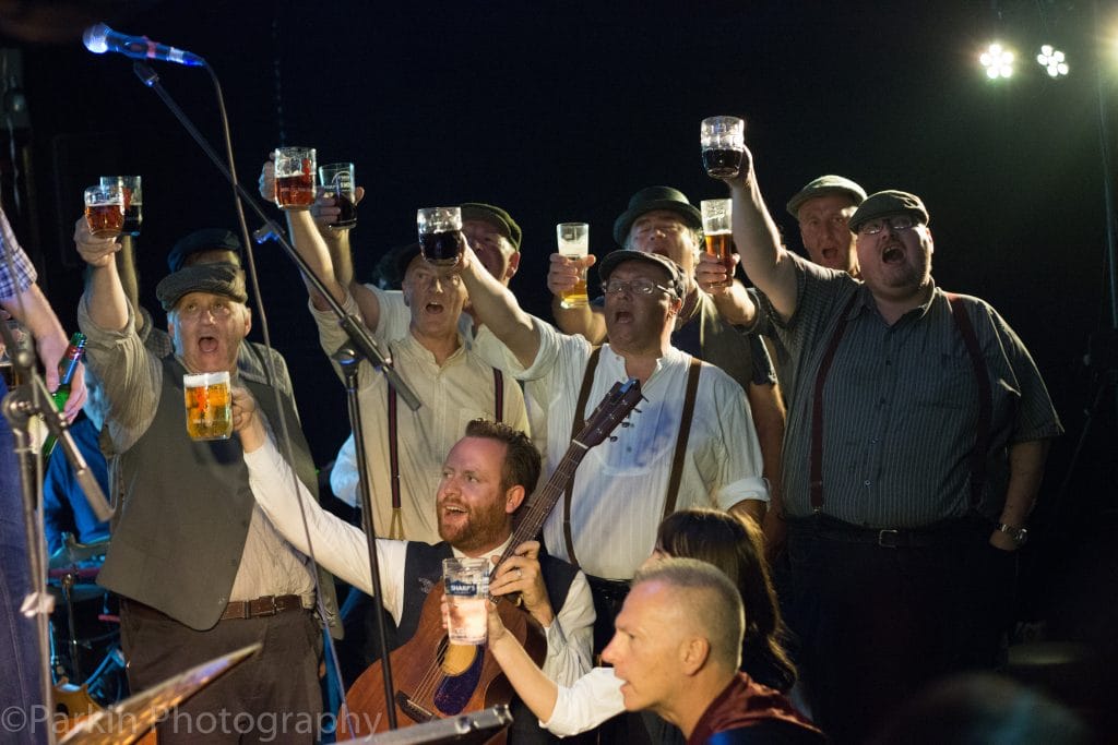 Image shows a group of men in white shirts, with braces and wearing flat caps look of into the distance, while raising their pints in glass tankards.