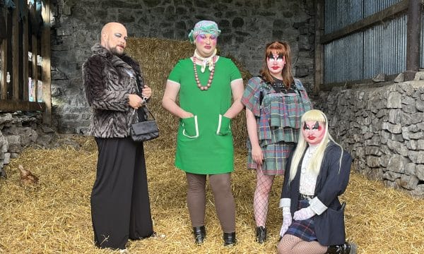 Four performers in drag, three standing and one kneeling on the floor of the straw filled barn they're in.