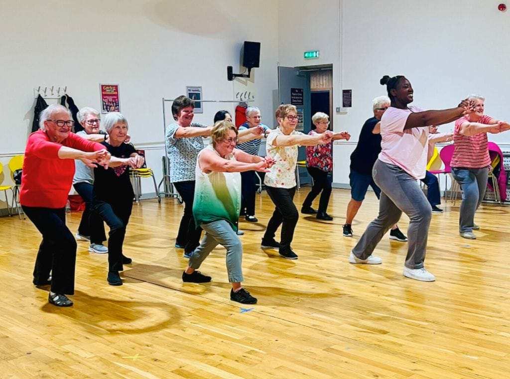 A group of people in a dance studio, they are all doing a lunge with their arms stretched out in front of them.