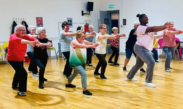 A group of people in a dance studio, they are all doing a lunge with their arms stretched out in front of them.