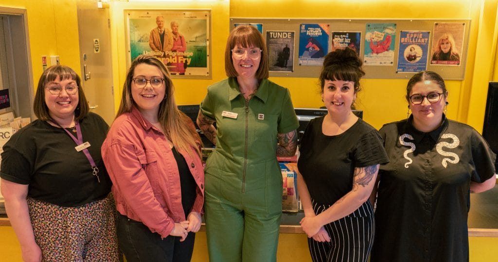 A photo of five young white women smiling, standing in front of a bright yellow reception area with posters on display.