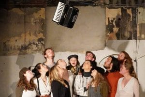 Twelve performers - eleven looking up at an accordion that has been thrown up into the air, and one looking directly at the camera with a surprised face