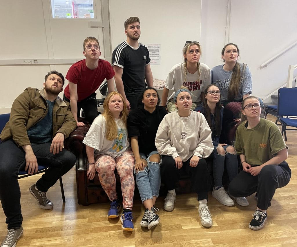 A cast of 10 students (most seated, some standing) in rehearsals for the show - they are all looking upwards in the same direction at something out of shot.