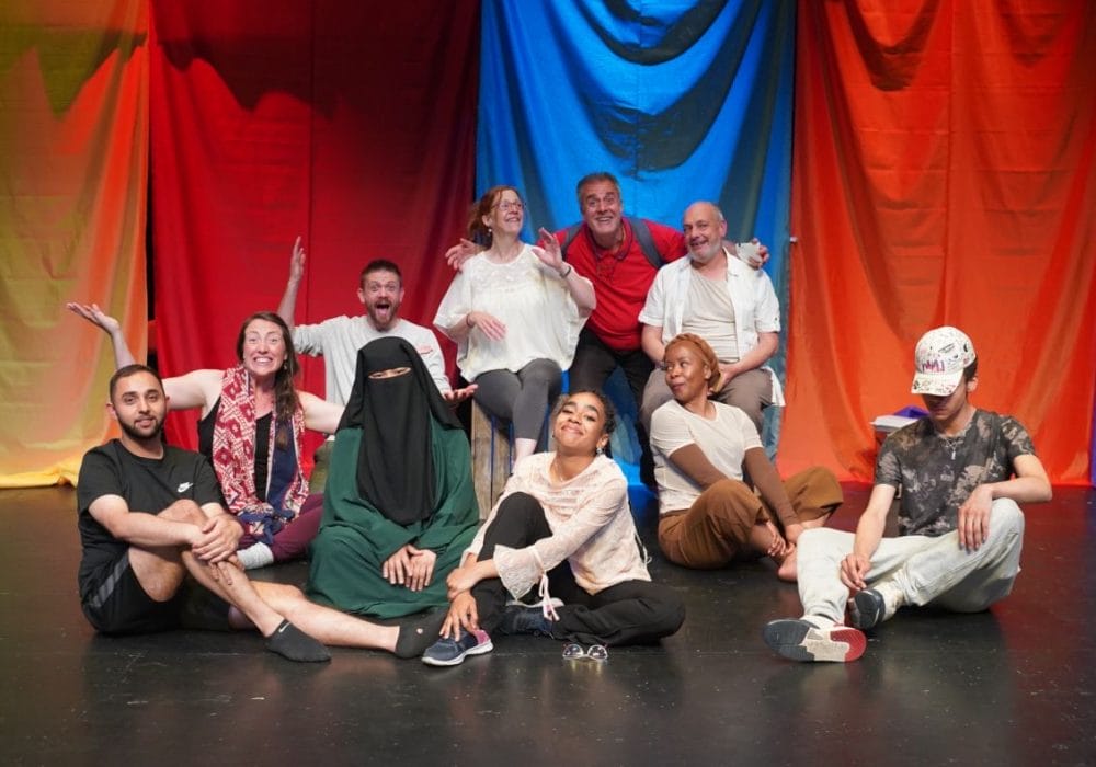 Alt: image of a group of people of various ethnicities sat on a theatre stage smiling at the camera, in the background there are number of colourful cloth banners hanging down.