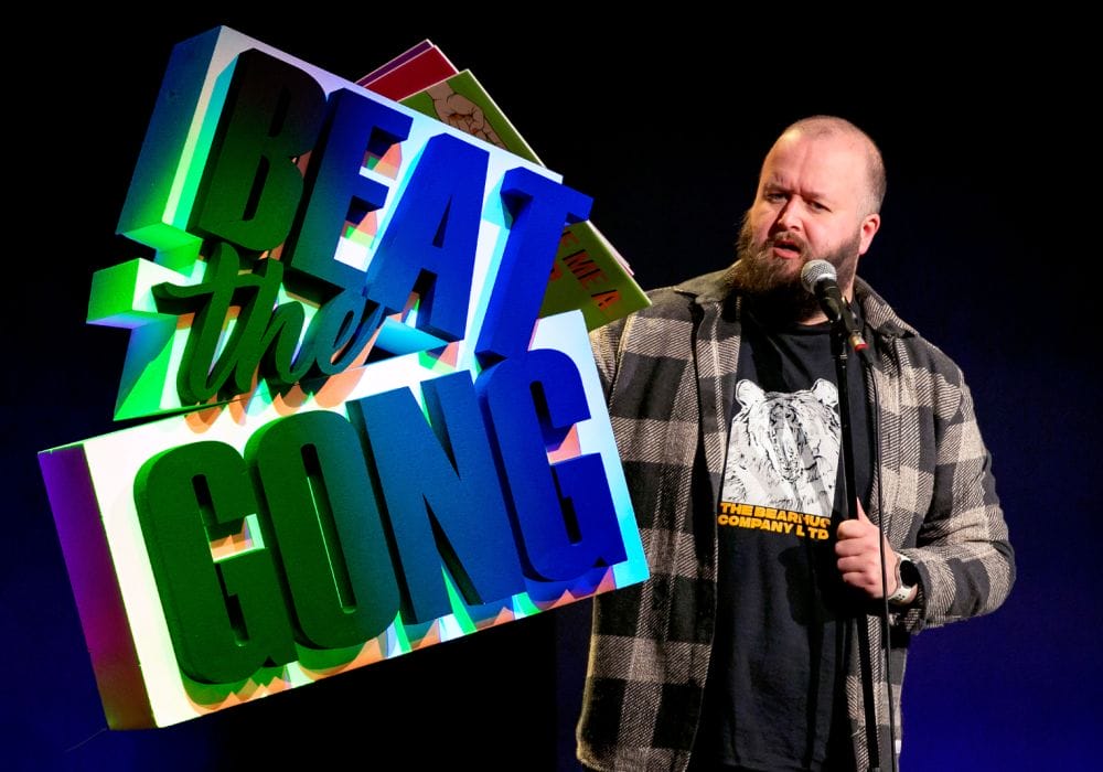 Comedian Freddy Quinne standing to the right of an illuminated Beat the Gong logo.