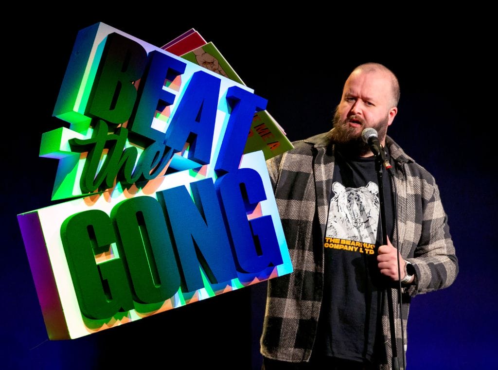 Comedian Freddy Quinne standing to the right of an illuminated Beat the Gong logo.