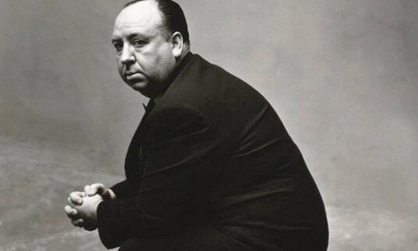 Director Alfred Hitchcock sits side-on, he is looking towards the camera. The photo is in black and white.