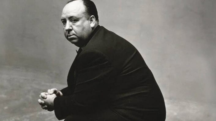 Director Alfred Hitchcock sits side-on, he is looking towards the camera. The photo is in black and white.