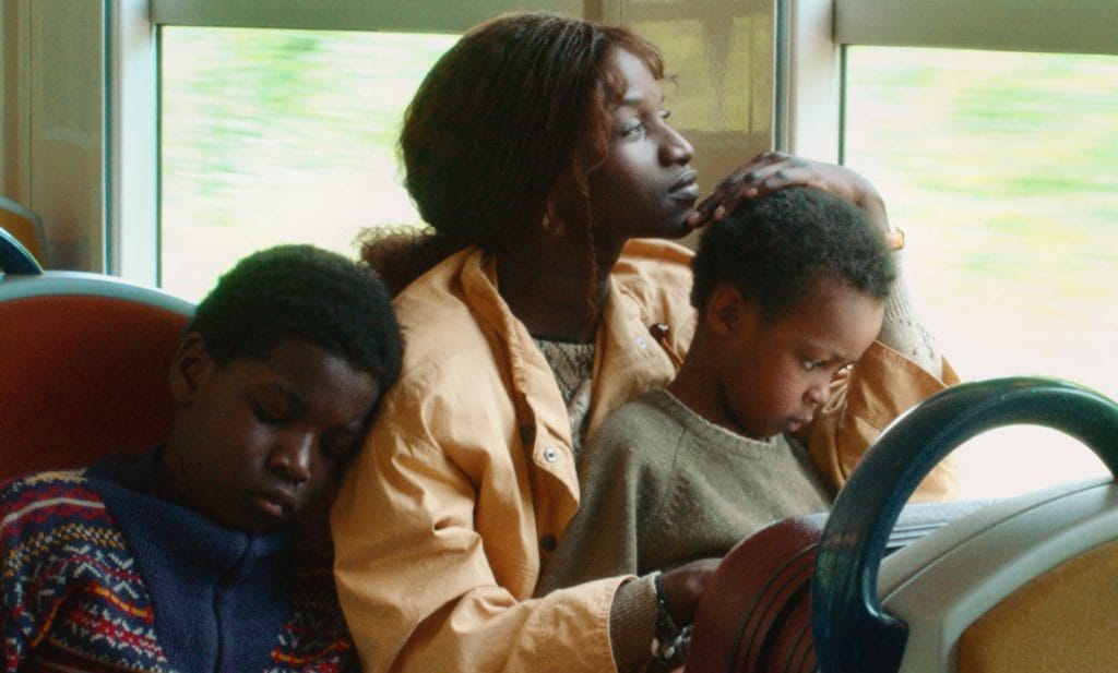 A mother sits on a bus, one child on her lap and another sitting next to her with their head on her shoulder