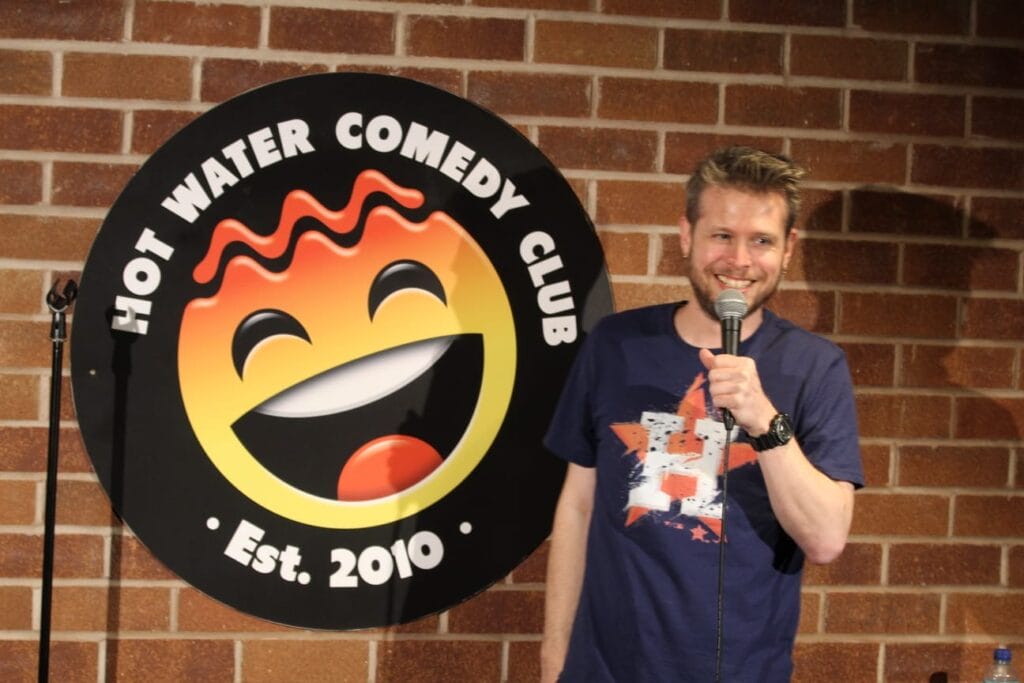 Comedian Phil Chapman, a white man with short brown hair, is holding a microphone and standing in front of the Hot Water Comedy Club logo. He is grinning broadly.