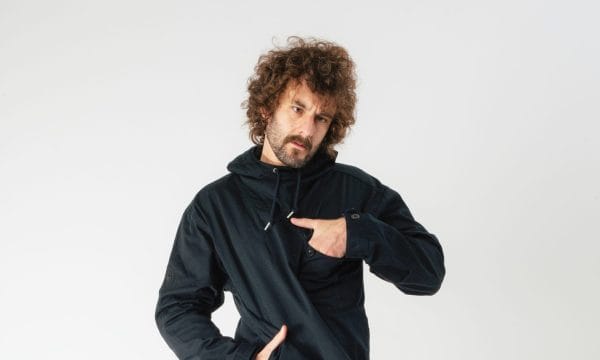 Comedian Josh Pugh, he is standing in front of a white background with one hand in his pocket on his waist, and the other hand in a pocket on his chest.