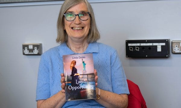 Creative Writing participant and published author Linda Dooks. Linda is a white woman, with shoulder length blonde-grey hair. She is holding a copy of her newly published book, and is smiling proudly.