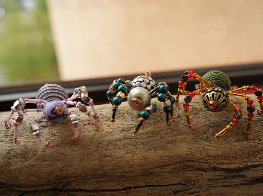 Three multi-coloured spiders made of beads standing on a piece of driftwood.