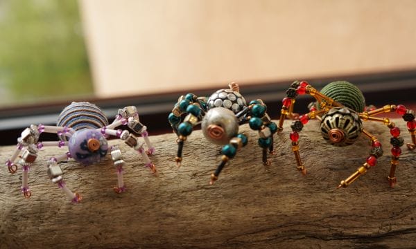 Three multi-coloured spiders made of beads standing on a piece of driftwood.