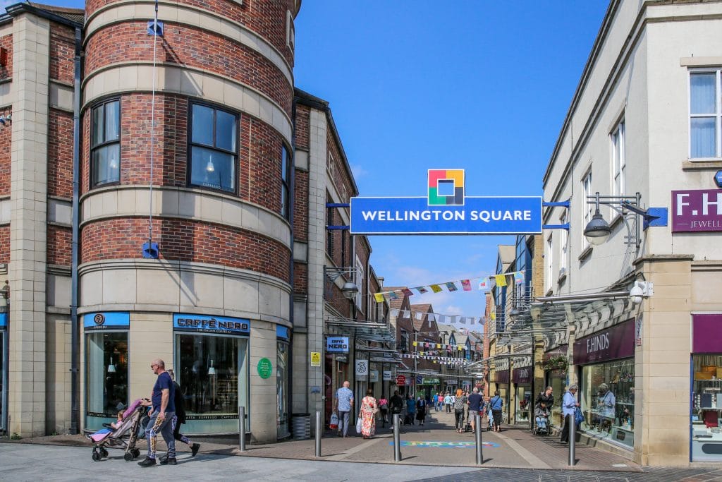 A shot looking down the lane of outdoor shopping centre, Wellington Square, in Stockton. It's a sunny day with a bright blue sky, and there is bunting strung between the buildings on the two sides of the lane. Plenty of people are out shopping.